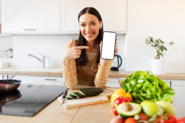 Harika bir aşçılık uygulaması. Neşeli bayan yemek pişirirken boş akıllı telefon ekranını işaret ediyor ve reklam veya web sitesi öneriyor.