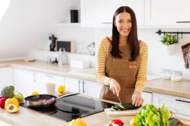 Mutlu bayan sebze salatası hazırlıyor, salatalıkları kesiyor, açık mutfakta duruyor ve kameraya gülümsüyor.