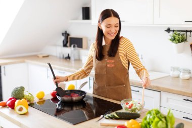 Genç bir kadın mutfakta yemek pişiriyor, birden fazla iş yapıyor, tavada et pişiriyor ve kasede sebze salatası pişiriyor, akşam yemeğini hazırlıyor.