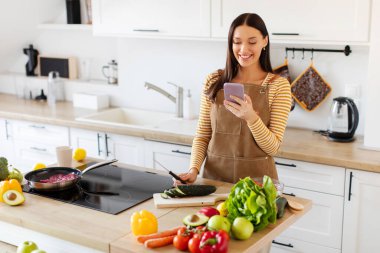 Mutlu kadın yemek hazırlarken, mutfak masasında sebze keserken ve internetten mesaj atarken ya da marketten sipariş verirken akıllı telefon kullanıyor.