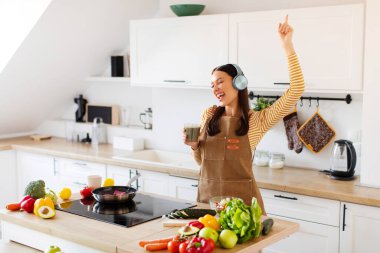Kablosuz kulaklıklı neşeli bayan, masada yemek pişirirken dans ediyor ve smoothie ile bardak tutuyor.