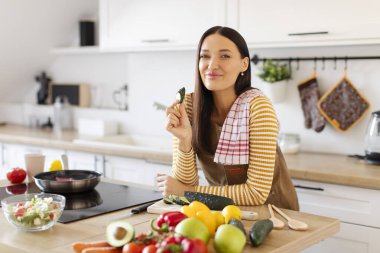 Mutlu, güzel bir kadın yemek hazırlıyor ve bir dilim salatalık yiyor. Sıcak bir mutfakta kameraya gülümsüyor.