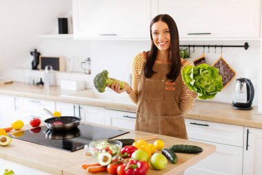 Brokoli ve marul tutan, salata için ürünler seçen, mutfakta bekleyen genç ve mutlu bir kadın. Sağlıklı gıda ve diyet konsepti