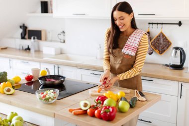 Modern mutfakta havuç soyan ve sağlıklı malzemelerle çevrili taze sebze salatası hazırlayan genç bir kadın.