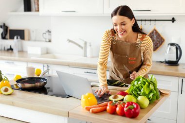 Çevrimiçi aşçılık dersi. Vloglama için dizüstü bilgisayar kullanan bir kadın, sebzeli vejetaryen salata yemeği pişirirken yemek için yemek tarifi arayan bir kadın.