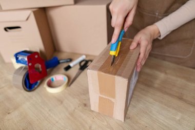 Karton kutuyu açarken ofis bıçağı kullanan kadın, masada duruyor ve paketleri açıyor, yakın plan.