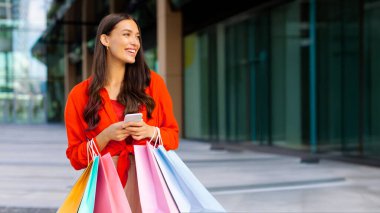 Bir sürü çantası olan mutlu Avrupalı kadın akıllı telefon kullanıyor, online taksi çağırıyor, alışveriş merkezinin önünde dikiliyor. E-ticaret kavramı