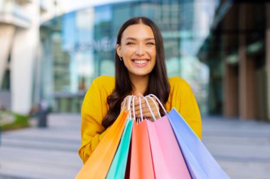 Alışveriş meraklısı heyecanlı bir kadın alışveriş torbalarıyla dışarıda poz veriyor, hafta sonları veya mevsimsel satışların keyfini çıkarıyor. Ticaret ve indirimler