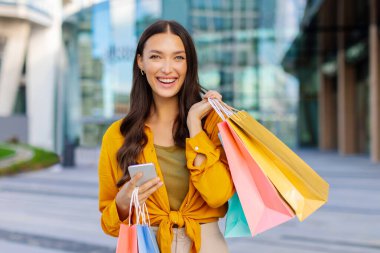 Büyük mevsimlik satışlar. Alışveriş çantaları ve akıllı telefonları olan mutlu bir kadın alışveriş merkezinin yakınındaki sokakta kameraya gülümsüyor.