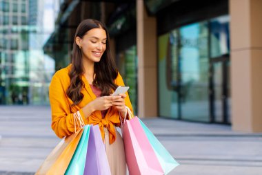 İndirim ile ilgili mesaj. Renkli alışveriş çantalarıyla neşeli genç bir kadın akıllı telefonuyla alışveriş merkezinin önünde yürüyor.