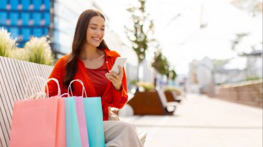 Alışveriş merkezinin dışında alışveriş torbalarıyla bankta otururken cep telefonu kullanan genç bir kadın.