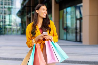Bir sürü çantası olan mutlu Avrupalı kadın akıllı telefon kullanıyor, online taksi çağırıyor, alışveriş merkezinin önünde dikiliyor. E-ticaret kavramı