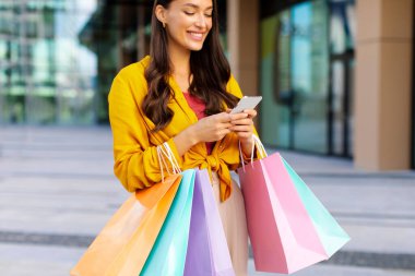 Alışveriş meraklısı kadın cep telefonunu açık havada kullanıyor. Alışveriş merkezinde başarılı bir şekilde alışveriş yaptıktan sonra bir sürü kese kağıdıyla yürüyor. Nakit bilgilerini kontrol ediyor.