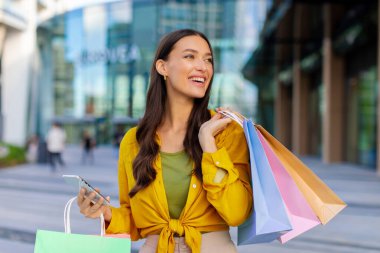 Hafta sonu ve alışveriş zamanı. Alışveriş merkezinin yakınında duran çantalı ve akıllı telefonlu mutlu genç bir kadın.