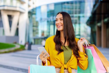 Güzel bayan çantalarla online alışveriş yapmaktan hoşlanıyor, alışveriş merkezinin yakınında cep telefonu kullanıyor ve başka yere bakıyor. İndirim ve satışlarla ilgili mesajlar için yeni mobil uygulama