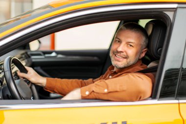 Yakışıklı olumlu erkek taksi şoförü kameraya bakıyor ve gülümsüyor, sürücü koltuğunda oturuyor, yolcu bekliyor, yan görüntü çekimi var.