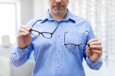 Erkek müşteri optik dükkanında yeni gözlük seçiyor, elinde iki çerçeve var. Görme bozukluğu ve görme bozukluğu.