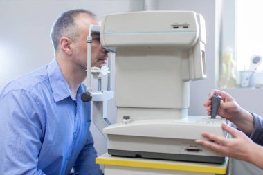Optometrist kabinesinde oturan bir adam görme yeteneğini profesyonel bir gözlükçünün özel ekipman makinesiyle test ediyor.