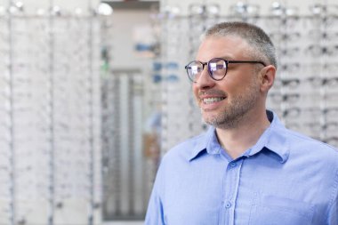 Görme ve görme konsepti. Gözlük takan mutlu, orta yaşlı bir adam, gözlüklü ve gözlüklü bir sürü gözlükçüye karşı optik mağazasında duruyor.
