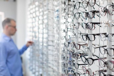 Bir optik mağazanın parlak duvarında çeşitli gözlükleri inceleyen orta yaşlı bir adam görüş düzeltmesi için yeni çerçeve seçiyor.