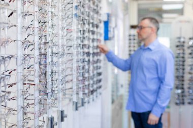 Erkek müşteri farklı gözlüklere bakıyor, optik mağazada yeni gözlük seçiyor ve deniyor, görüş düzeltmesi için kare seçiyor, gözlük rafına odaklanıyor