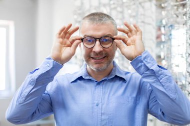 Oftalmoloji ve göz bakımı konsepti. Optik mağazasında yeni gözlük deneyen mutlu erkek müşteri ve kameraya gülümseyen adam çerçeveyi seçip seçiyor.