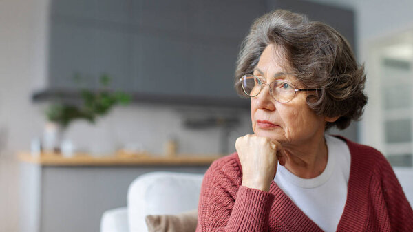 Thoughtful aged woman resting sitting on sofa, looking aside at free space, deep in memories and thoughts, panorama