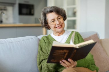 Kanepede oturup kitap okuyan, tasasız boş zamanlarının keyfini çıkaran mutlu Avrupalı yaşlı kadın.