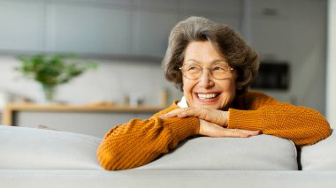 Mutlu, rahat, yaşlı bir kadın. Hayal kuruyor, kanepeye oturuyor, yan bakıyor ve gülümsüyor. Boş alanı olan bir panorama.