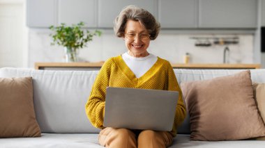 Gülümseyen yaşlı kadın dizüstü bilgisayar ekranına bakıyor, evdeki koltukta oturuyor, boş zamanlarını kanallarda geçiriyor, film seyrediyor, video görüşmesi yapıyor, panorama seyrediyor.