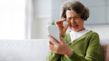 Yaşlı yaşta görme bozukluğu ve sağlık sorunları. Üst düzey bir kadın cep telefonu ekranına bakarken gözlerini kısıyor, evdeki koltukta oturuyor, fotokopi alanı olan bir panorama.