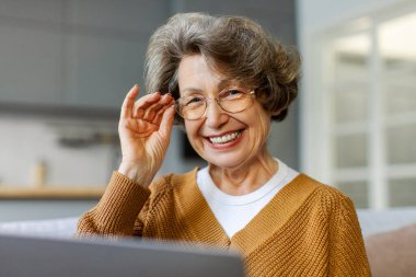 60 'lı yaşlarda dizüstü bilgisayar kullanan, haber okuyan sosyal medyaya göz atan, gözlüklerine dokunan ve kameraya gülümseyen yaşlı bir kadının portresi.