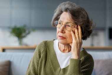 Kafasına dokunan mutsuz, baş ağrısı çeken, evde tek başına oturan, üzgün, düşünceli bir yüz ifadesiyle başka yere bakan yaşlı bir kadın.