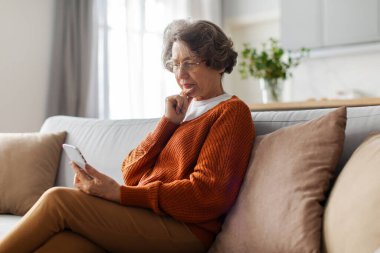Gözlüklü yaşlı kadın ciddi ve düşünceli bir yüz ifadesiyle telefon ekranına bakıyor, kötü haberler okuyor, evdeki kanepede oturuyor.