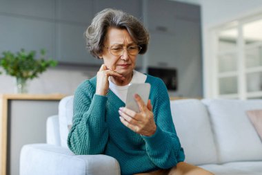 Düşünceli, üzgün, son sınıf öğrencisi bir kadın cep telefonu ekranına bakıyor, kötü haberler okuyor, online sohbet mesajı alıyor, kanepede oturuyor. Yaşlılık sorunları kavramı