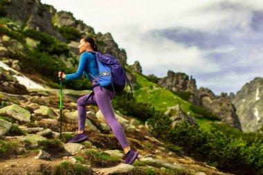 Güneşli bir yaz günü dağlarda yürüyüş yapan, sırt çantası takan ve yürüyüş direkleri kullanan, fotokopi çeken genç bir kadın.