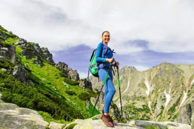 Sırt çantalı kadın gezgin dağların tepelerinde sopalarla yürüyüş yapıyor aktif yaşam tarzının ve güzel doğa manzarasının tadını çıkarıyor.