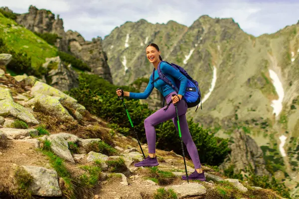 Sırt çantalı heyecanlı bayan dağların tepelerinde sopalarla yürüyor ve kameraya gülümsüyor, güzel manzaranın ve güneşli günün tadını çıkarıyor.