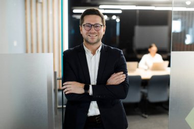 Ofis konferans salonunun dışında kollarını kavuşturup kameraya bakıp gülümseyen başarılı Avrupalı iş adamının portresi.