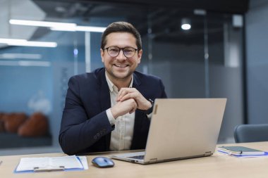 Ofiste masa başında oturan başarılı iş adamının portresi. Belgeler ve dizüstü bilgisayarla çalışıyor. Kameraya güvenle gülümsüyor.