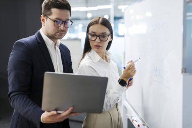 Ortaklık, takım çalışması. İş adamları dizüstü bilgisayarla proje üzerinde birlikte çalışıyor, beyaz tahtanın yanında duruyor, beyin fırtınası yapıyor, gelir tahmini yapıyorlar.