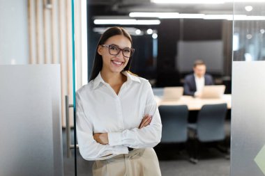 Profesyonel iş kadını portresi gülümsüyor, modern ofis ortamında kollarını kavuşturup poz veriyor, güven ve profesyonellik yayıyor.