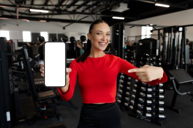 Mobil model. Boş akıllı telefon ekranını işaret eden kadın fitness reklamı ya da web sitesi için boş yer gösteriyor. Modern spor salonunda poz veriyor.