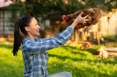 Kahverengi tavuk tutan ve gülümseyen genç bir çiftlik işçisi çiftlik arka bahçesinde poz veriyor.