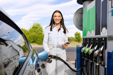 Heyecanlı genç bir kadın arabaya benzin dolduruyor, akıllı telefonu para ödemek için kullanıyor, kameraya bakıp gülümsüyor. Dükkanı ziyaret etmeden hızlı yakıt ikmali için mobil teknoloji kavramı