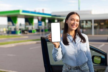 Çevrimiçi ECO yakıtı için cep telefonu görüntüsü veren mutlu genç bayan benzin istasyonunun yanında duruyor.