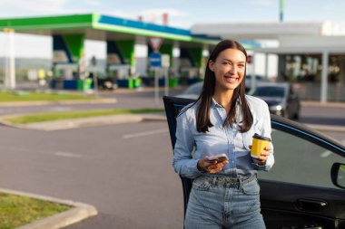 Benzin istasyonunda kahve içen genç bir kadın, arabasının yanında duruyor ve kameraya gülümsüyor, durup cep telefonu kullanıyor.