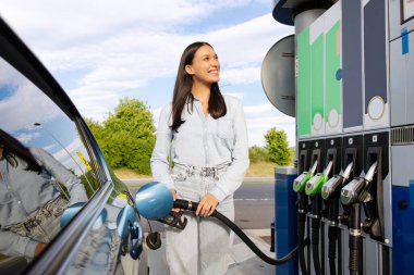 Benzin istasyonunda yakıt ikmali yapan bir kadın sürücü. Doldurma hortumunu tutuyor ve ekrana bakıyor.