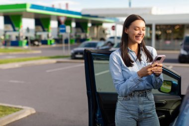 Benzin istasyonunun otoparkında cep telefonu kullanan mutlu, genç Avrupalı bir kadın.