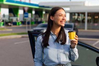 Benzin istasyonunda arabasının yanında kahve molası veren mutlu kadın sürücü, yolculuk sırasında mola veriyor, boş yer var.